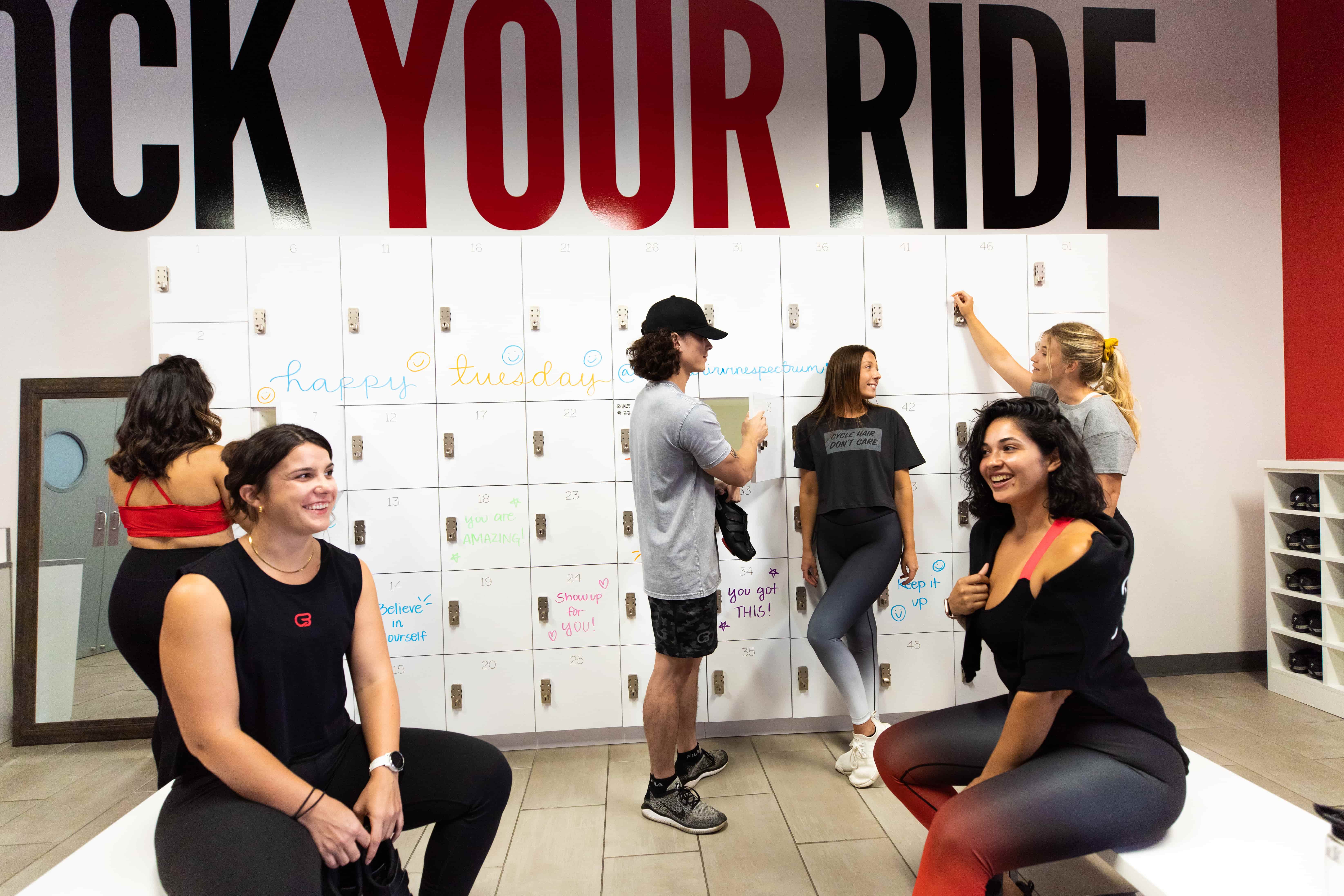 Individuals in a cycle bar locker room with lockers that have motivational writing for cycle beginners and experts